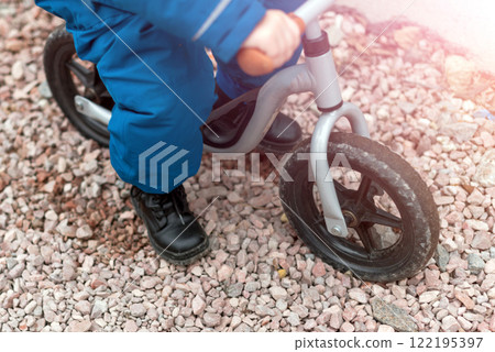 A young child riding a balance bike on a gravel pathway, wearing sturdy black shoes and blue pants. An active and playful moment, emphasizing outdoor activity, childhood exploration, and learning. A young child riding a balance bike on a gravel pathway, wearing sturdy black shoes and blue pants. An active and playful moment, emphasizing outdoor activity, childhood exploration, and learning. 122195397