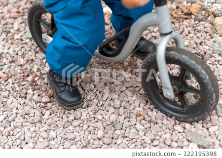 A young child riding a balance bike on a gravel pathway, wearing sturdy black shoes and blue pants. An active and playful moment, emphasizing outdoor activity, childhood exploration, and learning. 122195398