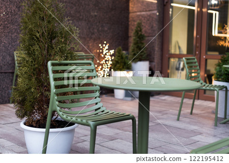 An inviting outdoor cafe area featuring green metal furniture, potted evergreens, and warm decorative lights. A serene and modern setting for relaxation and enjoying a peaceful urban atmosphere. 122195412