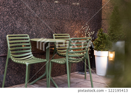 An inviting outdoor cafe area featuring green metal furniture, potted evergreens, and warm decorative lights. A serene and modern setting for relaxation and enjoying a peaceful urban atmosphere. 122195413