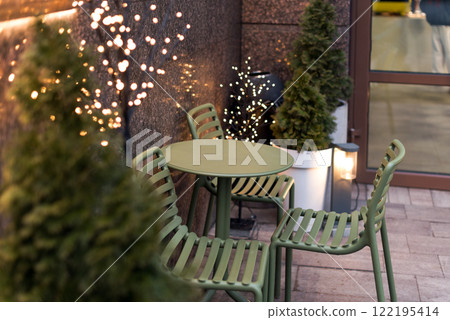 An inviting outdoor cafe area featuring green metal furniture, potted evergreens, and warm decorative lights. A serene and modern setting for relaxation and enjoying a peaceful urban atmosphere. An inviting outdoor cafe area featuring green metal furniture, potted evergreens, and warm decorative lights. A serene and modern setting for relaxation and enjoying a peaceful urban atmosphere. 122195414