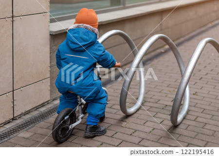 A young child dressed warmly explores an outdoor space while riding a small bike. The child interacts with an urban bike rack. Curiosity, activity, and discovery in a modern city environment. 122195416