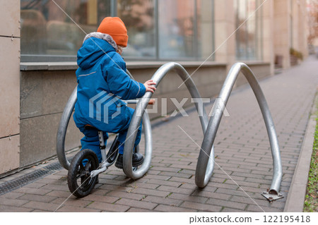 A young child dressed warmly explores an outdoor space while riding a small bike. The child interacts with an urban bike rack. Curiosity, activity, and discovery in a modern city environment. A young child dressed warmly explores an outdoor space while riding a small bike. The child interacts with an urban bike rack. Curiosity, activity, and discovery in a modern city environment. 122195418