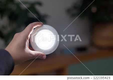 Hand Holding a Compact LED Light in a Dimly Lit Room Among Smooth Backgrounds. Innovation and portability of modern lighting solutions in everyday situations. Blackout concept. Hand Holding a Compact LED Light in a Dimly Lit Room Among Smooth Backgrounds. Innovation and portability of modern lighting solutions in everyday situations. Blackout concept. 122195423