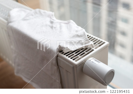A neatly spread white T-shirt placed on a home radiator, basking in sunlight streaming through a nearby window. Represents eco-friendly cloth drying and the utilization of home heating systems. 122195431