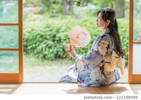 一位年輕女子坐在古民居的門廊上，用扇子搧風降溫 122196099