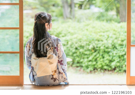 A young woman sitting on the veranda of an old house, cooling off by fanning herself with a paper fan 122196103