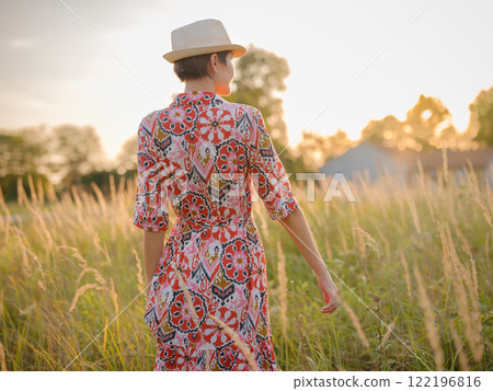 Young woman walking through picturesque European field in late summer. Golden sunlight, lush greenery, and serene rural atmosphere create peaceful countryside scene. Young woman walking through picturesque European field in late summer. Golden sunlight, lush greenery, and serene rural atmosphere create peaceful countryside scene. 122196816