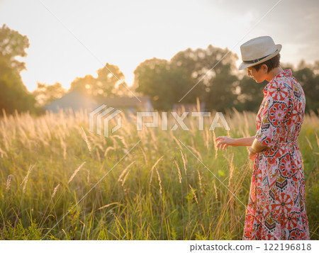 Young woman walking through picturesque European field in late summer. Golden sunlight, lush greenery, and serene rural atmosphere create peaceful countryside scene. 122196818
