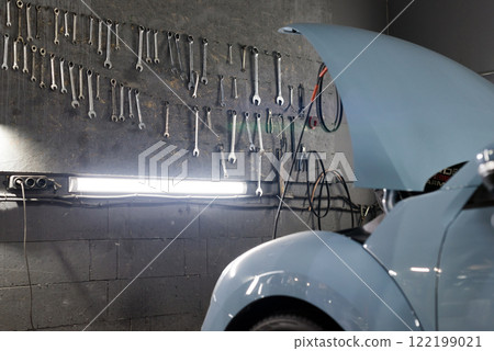 A mechanic garage with neatly arranged tool wall, a vehicle with the hood raised undergoing maintenance, and strong lighting. Technical expertise, automotive care, and repair efforts. 122199021