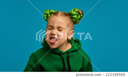 Child, girl in green hoodie, braided buns hairstyle, sticking out tongue playfully, symbolizing fun and carefree childhood against blue studio background 122199568