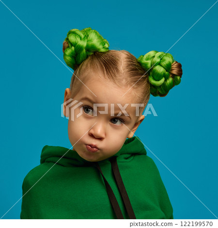 Girl, child puffing cheeks in thoughtful pose, wearing green hoodie, braided buns hairstyle, symbolizing curiosity and imagination against blue studio background 122199570