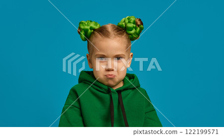 Preschool girl with pouty expression, in green hoodie, braided buns hairstyle, showing dissatisfaction and contemplation against blue studio background 122199571