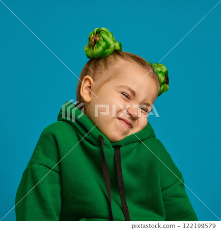 Girl, child with playful mischief, scrunched face, wearing green hoodie, braided buns hairstyle against blue studio background 122199579