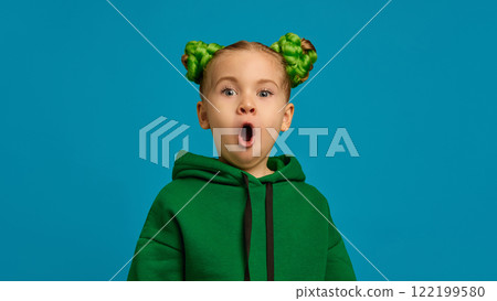 Portrait of cute little girl, child in green hoodie and braid buns hairstyle, expressing positive excitement and shock against blue studio background 122199580