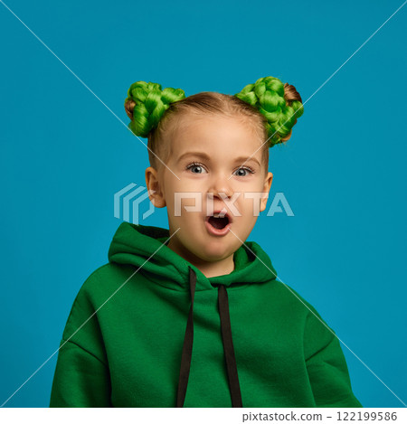 Portrait of emotional girl, child in casual green sweatshirt with stylish buns standing with open mouth, showing shock and excitement against blue studio background Portrait of emotional girl, child in casual green sweatshirt with stylish buns standing with open mouth, showing shock and excitement against blue studio background 122199586
