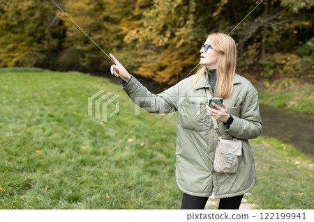 Hiker blonde woman holding smart phone and pointing finger something near river. Travel, technology and relax concept. High quality photo 122199941