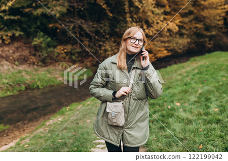 Stylish happy smiling modern young woman talking on phone, standing in autumn park, 30 girl holding smartphone on nature background. Female relaxing outdoors. High quality photo 122199942