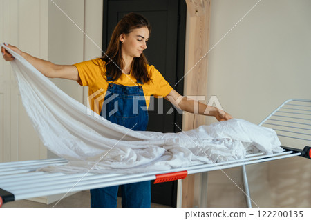 Woman wearing yellow t-shirt and blue jeans overalls Hanging Laundry On Drying Rack In Room. Everyday domestic life, highlighting productivity and routine activities concept. Woman wearing yellow t-shirt and blue jeans overalls Hanging Laundry On Drying Rack In Room. Everyday domestic life, highlighting productivity and routine activities concept. 122200135