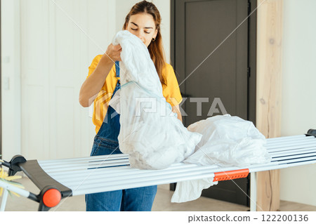 Beautiful young woman housewife wearing yellow t-shirt drying clothes on the clothesline after washing at home. Activity of daily living at house concept. 122200136
