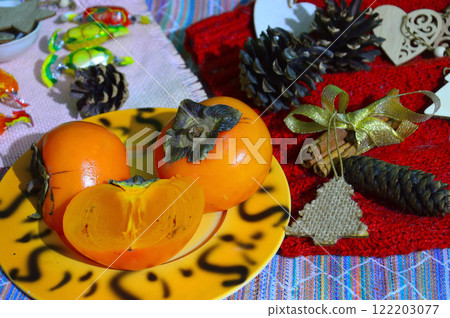 Still life of ripe bright persimmons, pine cones and candies on a crimson woolen background. Fruits lie on a beautiful plate. Whole and cut fruits. Still life of ripe bright persimmons, pine cones and candies on a crimson woolen background. Fruits lie on a beautiful plate. Whole and cut fruits. 122203077