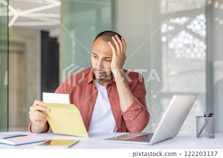 Man sitting at desk with a laptop, holding an envelope and looking concerned. Man sitting at desk with a laptop, holding an envelope and looking concerned. 122203137