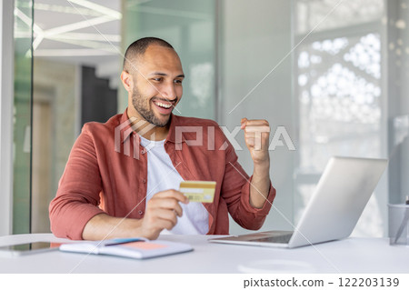 A cheerful man celebrates a successful online transaction by utilizing a credit card and laptop in a bright office setting. The image conveys concepts of digital technology, satisfaction. A cheerful man celebrates a successful online transaction by utilizing a credit card and laptop in a bright office setting. The image conveys concepts of digital technology, satisfaction. 122203139