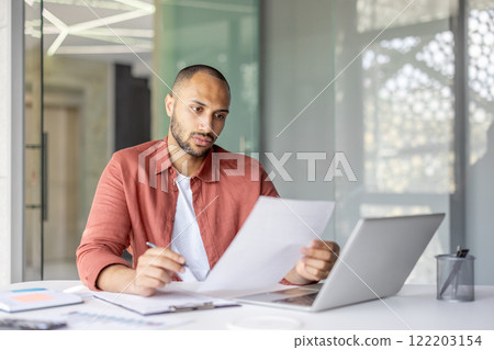 Business person analyzing paperwork on desk with laptop in a contemporary office environment, demonstrating focus and productivity. Business person analyzing paperwork on desk with laptop in a contemporary office environment, demonstrating focus and productivity. 122203154