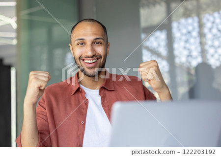 A cheerful individual in an office environment expressing joy with raised fists, representing success and positivity. The light-filled background complements sense of accomplishment conveyed. 122203192