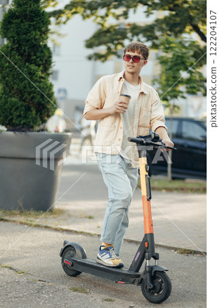 Smiling young student man with take away coffee going to start with sharing service kick- scooter in the morning city 122204107