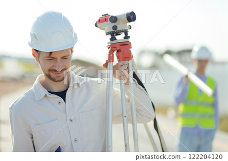 Engineer Surveyor Carries Tripod Level on shoulder. Walks at Construction Place Engineer Surveyor Carries Tripod Level on shoulder. Walks at Construction Place 122204120