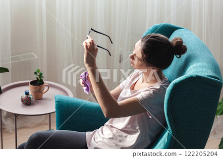 Young woman cleaning glasses with a napkin while sitting in a ch Young woman cleaning glasses with a napkin while sitting in a ch 122205204