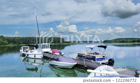There are several yachts in the water near the pier. The water in the lake is calm. There are several yachts in the water near the pier. The water in the lake is calm. 122206772