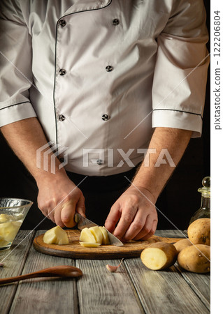 A skilled chef expertly chops potatoes on a wooden cutting board surrounded by fresh produce. The kitchen exudes a cozy atmosphere, enhanced by warm lighting and rustic decor A skilled chef expertly chops potatoes on a wooden cutting board surrounded by fresh produce. The kitchen exudes a cozy atmosphere, enhanced by warm lighting and rustic decor 122206804