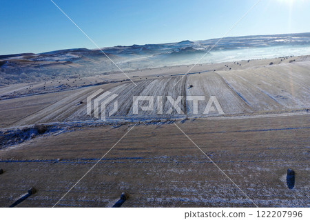 Aerial view of snow-covered field dotted with hay bales under a clear blue sky during winter Aerial view of snow-covered field dotted with hay bales under a clear blue sky during winter 122207996
