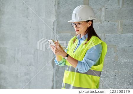Female industrial worker in protective vest helmet using smartphone at construction site Female industrial worker in protective vest helmet using smartphone at construction site 122208179