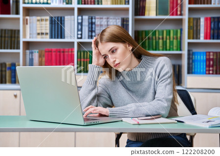 Sad teenage student girl preparing in classroom library looking at laptop 122208200