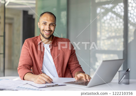Portrait of a young successful businessman financier at his workplace inside the office, the employee is smiling and looking at the camera, writing down data on a paper report document 122210356