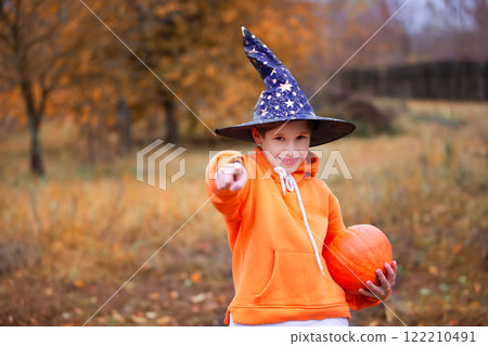 A child in an orange costume and a wizard hat for Halloween. A boy holds a pumpkin on an autumn background. A child at a Halloween party. 122210491