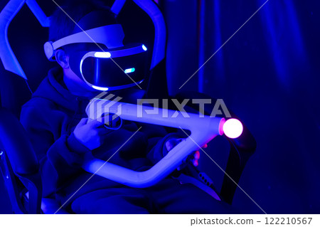 A boy is playing a video game with a controller. The controller is white and has a red light on it. The room is dark and the boy is sitting in a chair 122210567