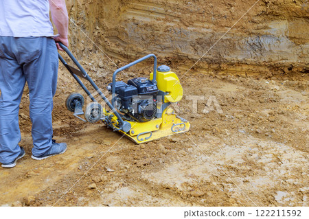 Contractor operates plate compactor on construction site, compacting soil for solid foundation. 122211592