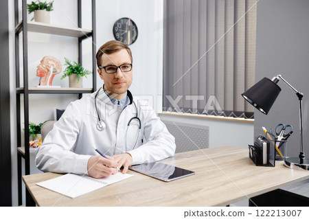 Portrait of young doctor wearing glasses and white uniform with stethoscope, speaking and consulting patient online, looking at camera, making video call, sitting at table in office Portrait of young doctor wearing glasses and white uniform with stethoscope, speaking and consulting patient online, looking at camera, making video call, sitting at table in office 122213007