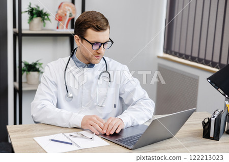 Young attractive otolaryngologist doctor sitting at his workplace in the office and holding a nose mirror Young attractive otolaryngologist doctor sitting at his workplace in the office and holding a nose mirror 122213023
