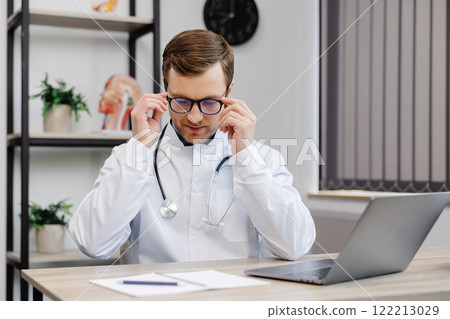 A young attractive doctor sits at his workplace in the office and takes off his glasses at the end of the working day A young attractive doctor sits at his workplace in the office and takes off his glasses at the end of the working day 122213029