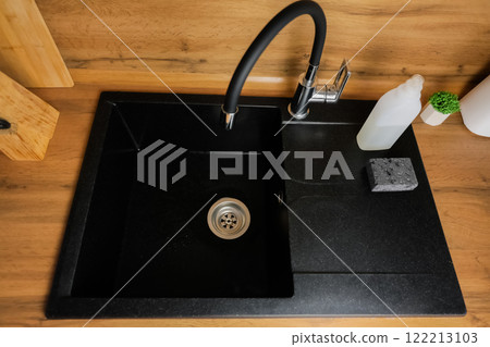 Black kitchen sink with flexible black faucet on wooden background, white detergent and black dishwashing sponge on the sink. The concept of washing dishes, modern cuisine, cleanliness. High quality 122213103