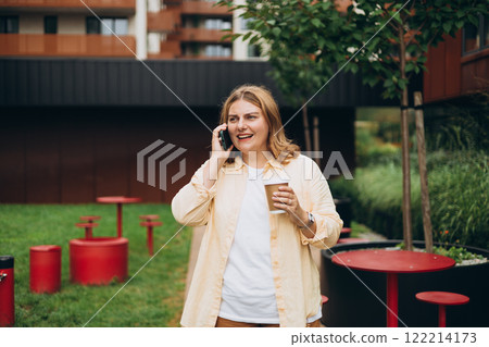 Beautiful smiling 30s female with coffee cup using smartphone and standing outdoors. Phone Communication. Happy cheerful young woman walking on city park, Urban lifestyle concept. Beautiful smiling 30s female with coffee cup using smartphone and standing outdoors. Phone Communication. Happy cheerful young woman walking on city park, Urban lifestyle concept. 122214173