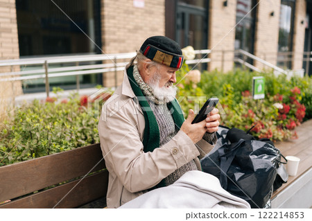 Side view of aged beggar man wrapped in blanket using smartphone sitting on urban bench outside building on cold autumn day. Concept of poverty and vulnerability, evoking empathy and compassion. Side view of aged beggar man wrapped in blanket using smartphone sitting on urban bench outside building on cold autumn day. Concept of poverty and vulnerability, evoking empathy and compassion. 122214853