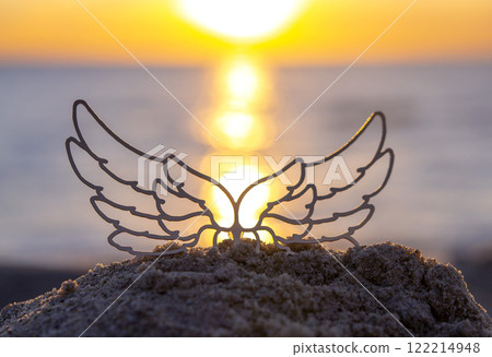 Black silhouette stick with transparent wings in sand on sandy beach of sea shore against sea background, sea waves and sun path during sunrise or sunset with rising or setting sun close up. Concept Black silhouette stick with transparent wings in sand on sandy beach of sea shore against sea background, sea waves and sun path during sunrise or sunset with rising or setting sun close up. Concept 122214948