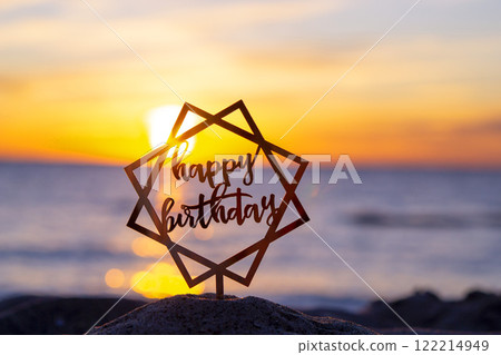 Black silhouette of stick with the words Happy Birthday in rectangular frame on background of sea and sea waves during sunset sunrise with sunrise or setting sun close-up. Concept holiday celebration 122214949