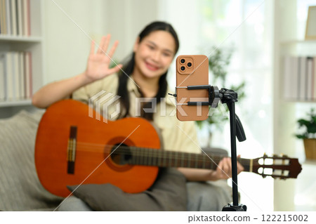 Cheerful woman playing guitar and waving at the camera during a live stream or video recording session 122215002